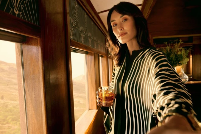Woman in striped top holds drink in vintage train with scenic landscape view, capturing a blend of elegance and travel ambiance.