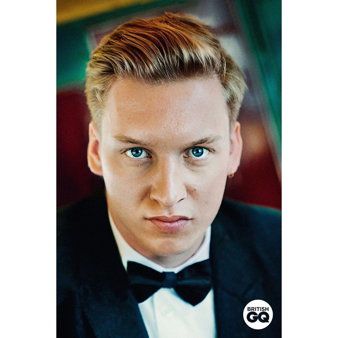 Close-up studio portrait of a young adult male with blonde hair and blue eyes, wearing a black tuxedo and bow tie, posing indoors.