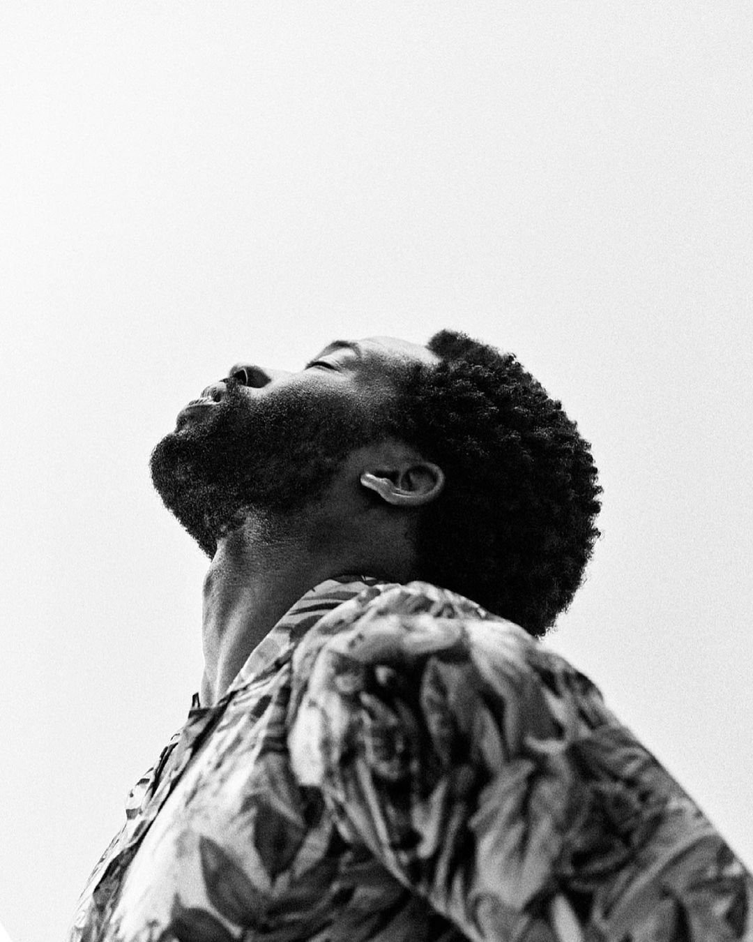Black man with short textured hair and beard looking upward wearing a patterned shirt against a pale sky background