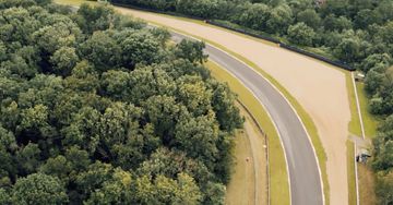 Aerial view of a racetrack winding through a dense green forest, showcasing its asphalt surface and surrounding natural landscape.