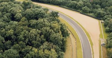 Aerial view of a racetrack winding through a dense green forest, showcasing its asphalt surface and surrounding natural landscape.