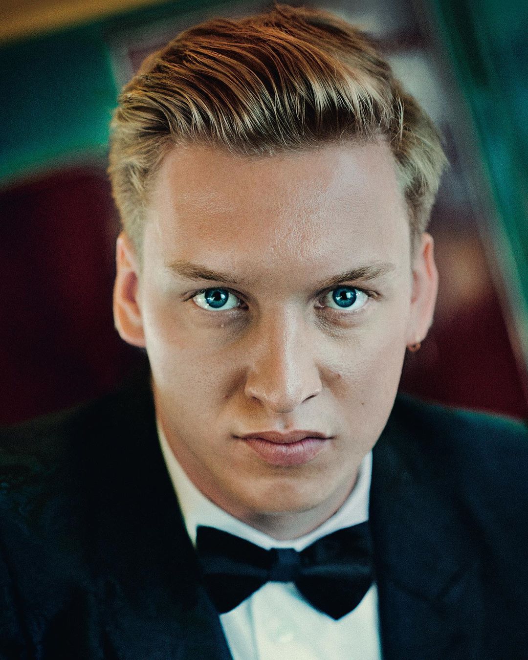 Close-up portrait of a blond adult male wearing a black bow tie and tuxedo-style jacket against a softly blurred background.