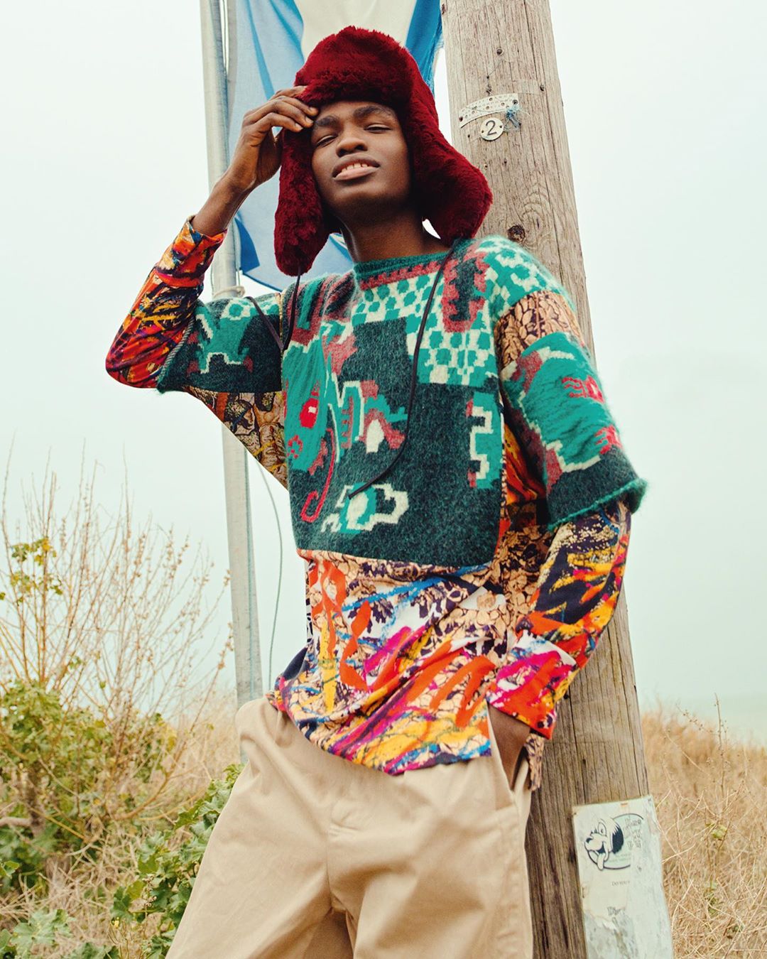 Young Black man in colorful, patterned sweater and fur hat stands outdoors by pole in fashion editorial style, looking stylish.