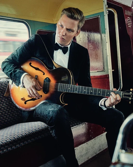 Blond man in a patterned tuxedo and bow tie sits playing a vintage guitar on a train with paneled walls and herringbone seats.