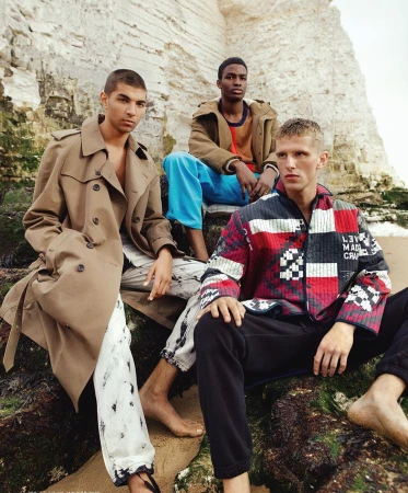 Three young adult males pose on rocks at a beach, wearing fashionable jackets and pants, with a white cliff face in the background.