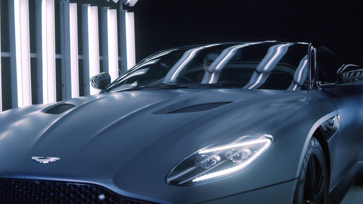 Close-up of a matte blue Aston Martin DBS Superleggera Volante with bright headlights in a studio setting, showcasing automotive design.