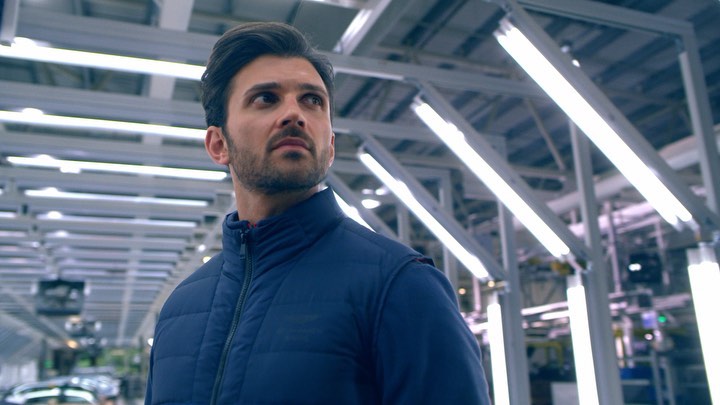 Adult male in a blue jacket looking up in an industrial facility with bright fluorescent lighting. The setting appears to be a factory.