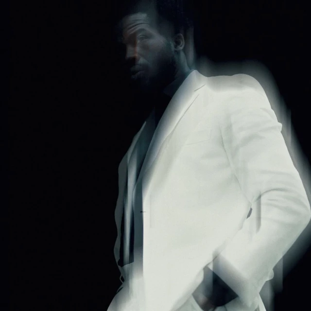 An adult male with dark skin in a light-colored suit stands against a dark background with a motion blur effect and diffused lighting.