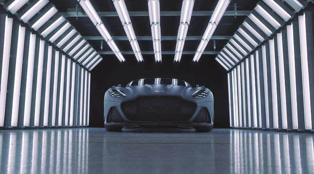 Sleek matte gray Aston Martin sports car showcased indoors with bright white fluorescent lighting creating a futuristic tunnel effect.