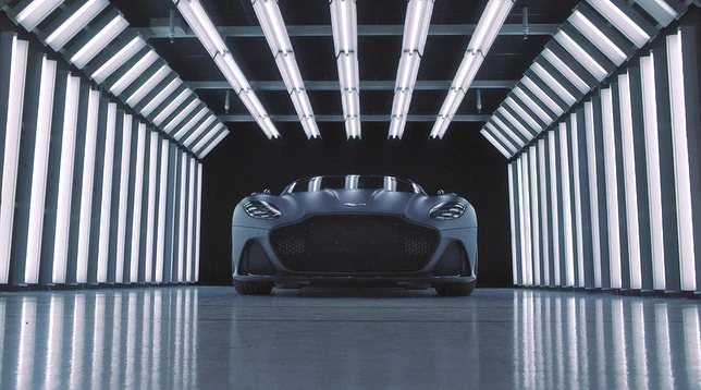 Sleek matte gray Aston Martin sports car showcased indoors with bright white fluorescent lighting creating a futuristic tunnel effect.