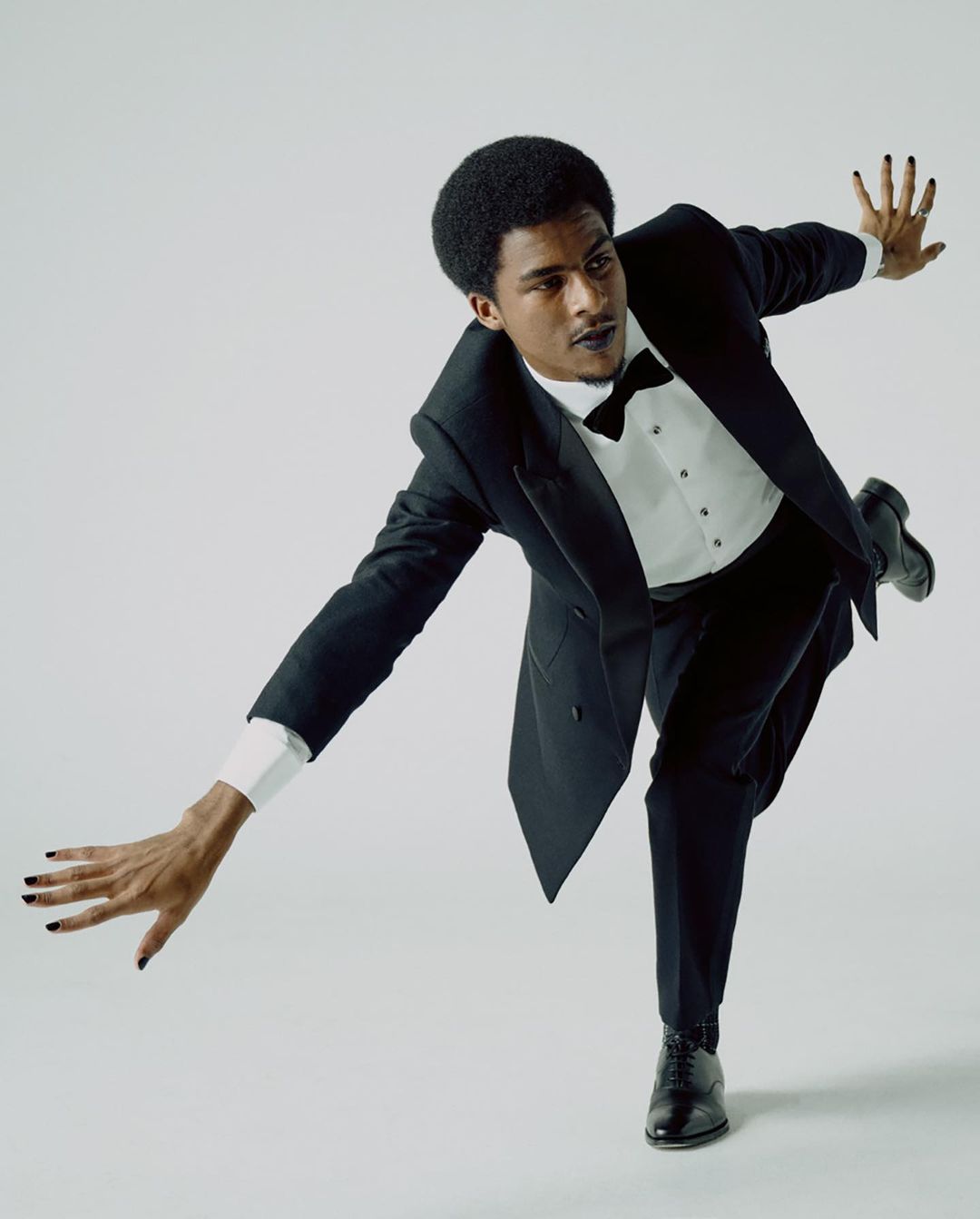 An African-American male model in a black tuxedo and polished black shoes posing dynamically in a studio with a light gray background.