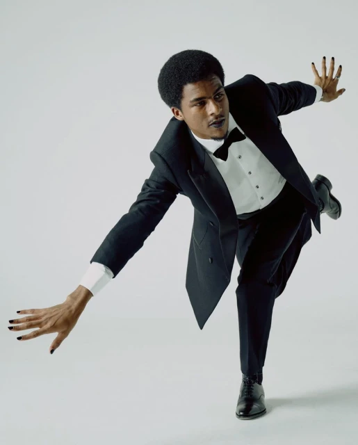 An African-American male model in a black tuxedo and polished black shoes posing dynamically in a studio with a light gray background.