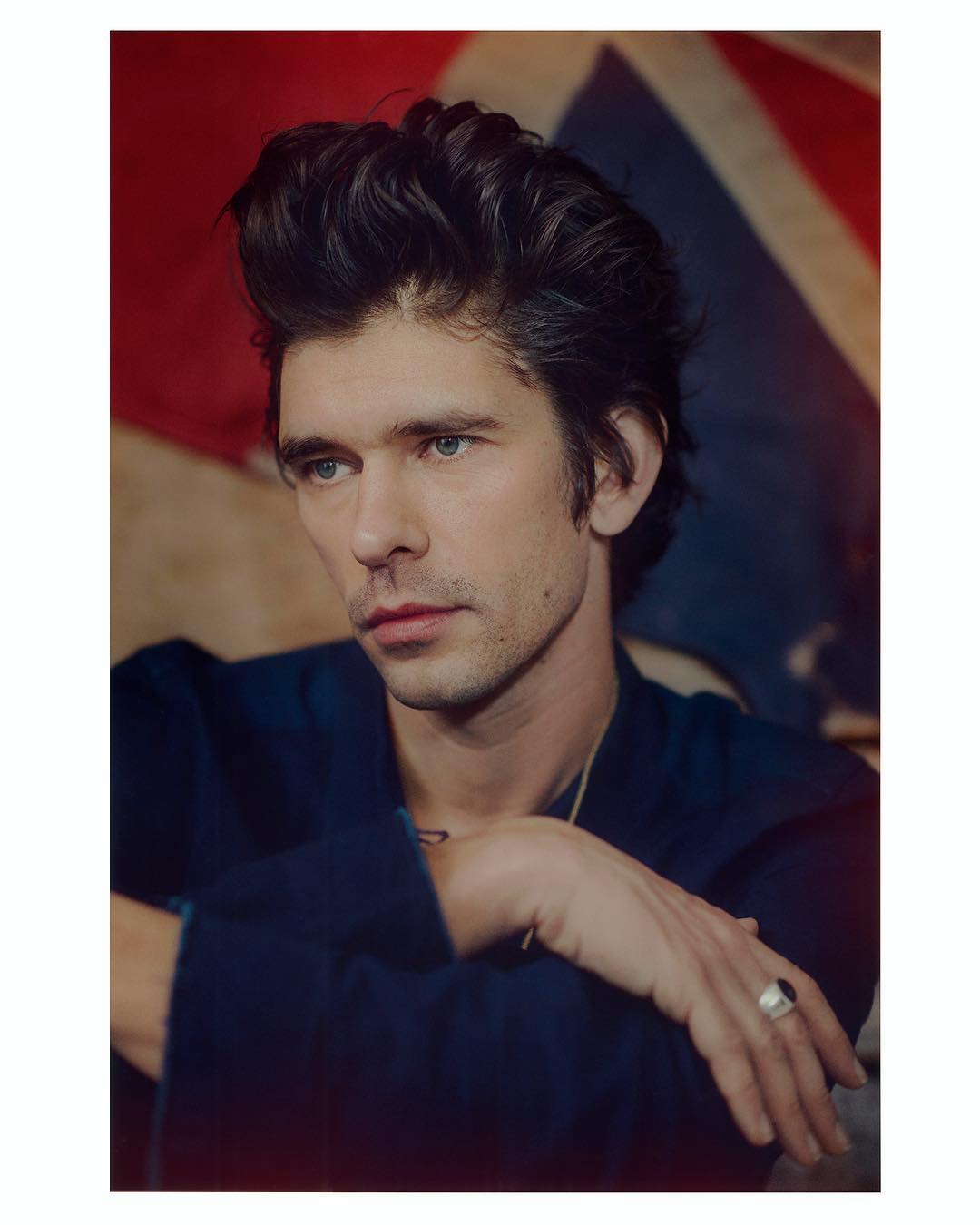 Portrait of a handsome adult man with dark hair and blue eyes, wearing a navy robe, posing in front of a British flag.