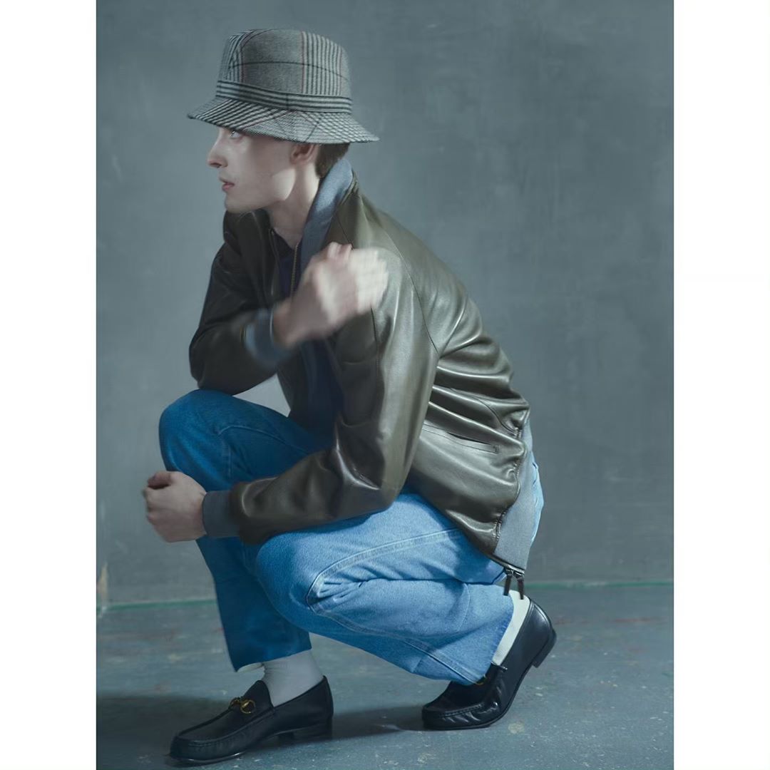 Young adult male in olive bomber jacket, blue jeans, and plaid bucket hat squats in a studio with a gray background and black loafers.