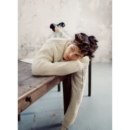 Young man in a cream knitted sweater lying prone on a wooden table with his arms resting over the edge.