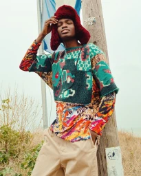 Man wearing a red furry trapper hat and patterned sweater leaning against a wooden pole outdoors.