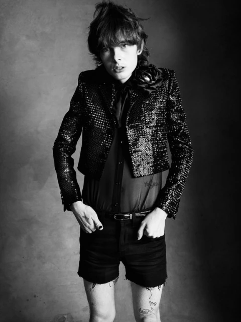 Black and white portrait of a stylish young adult male in a sequined jacket with a floral embellishment, posed against a dark backdrop.