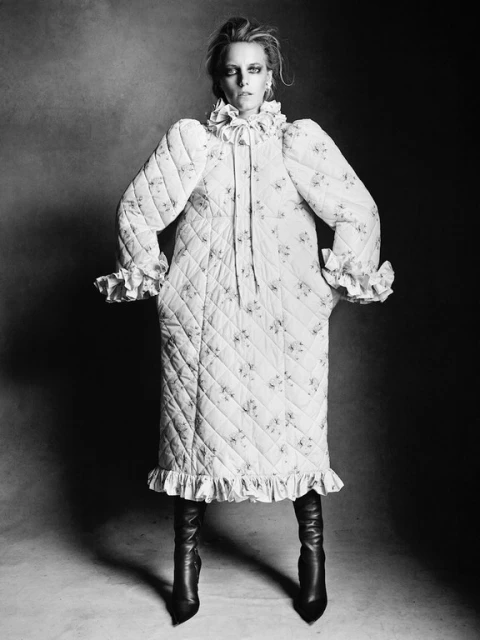 Black-and-white portrait of a woman in a floral quilted coat and black boots, posing against a dark textured backdrop in the photo studio.