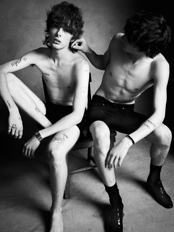 Two shirtless young men sit in a black-and-white portrait, one adjusting the other's hair, showcasing tattoos and intimate male bonding.