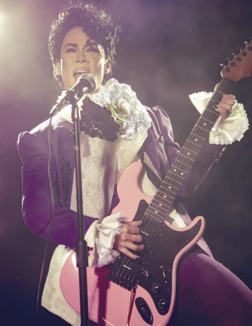 Singer in purple suit with ruffled collar and electric guitar performing on stage in spotlight with dark background and smoky haze.