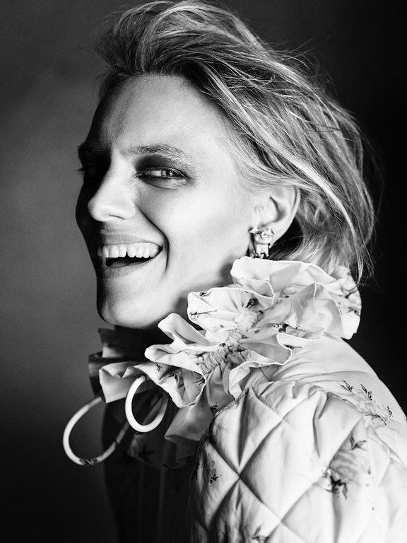 Monochrome portrait of a smiling adult female with short hair, wearing statement earrings and a quilted jacket with ruffled floral trim.
