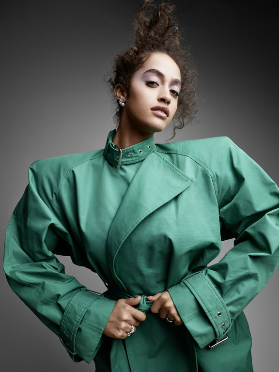 Model with voluminous curly hair wearing a structured emerald green trench coat against a plain grey background