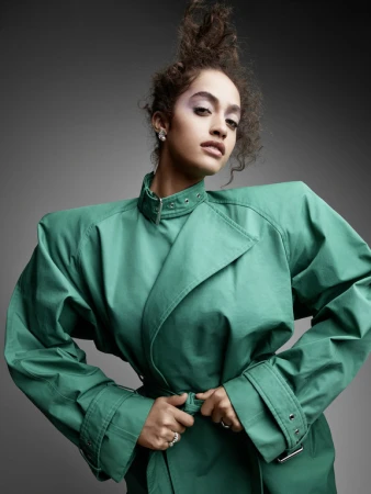 Model with voluminous curly hair wearing a structured emerald green trench coat against a plain grey background
