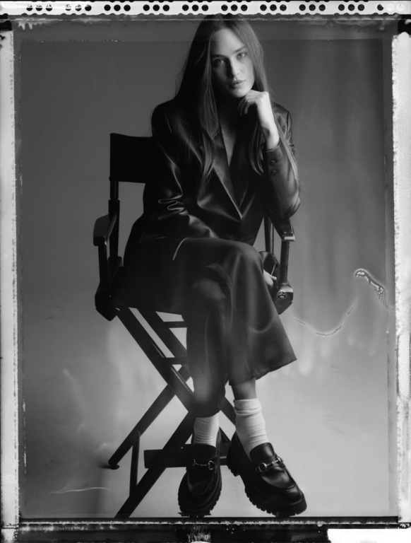 Monochrome portrait of a woman in a suit sitting on a director's chair, looking at the camera with a neutral expression in a studio.