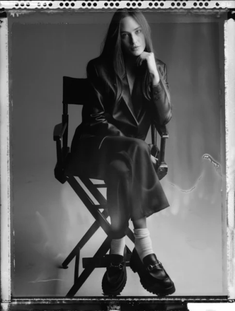 Monochrome portrait of a woman in a suit sitting on a director's chair, looking at the camera with a neutral expression in a studio.