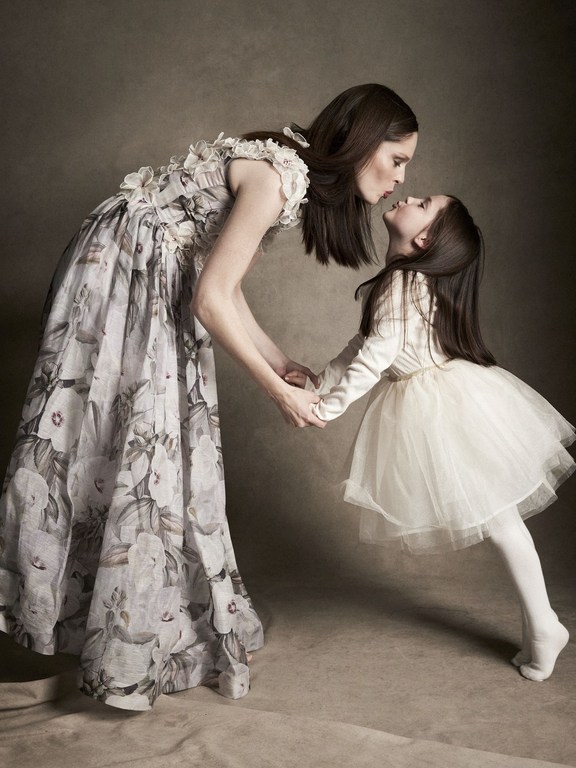 A mother and her young daughter face each other and hold hands in a studio portrait, both wearing white dresses in front of a brown backdrop.