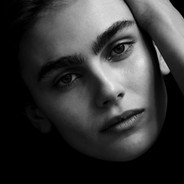 Close-up portrait of a young adult with light skin and dark hair against a black background, emphasizing facial features and natural skin texture.