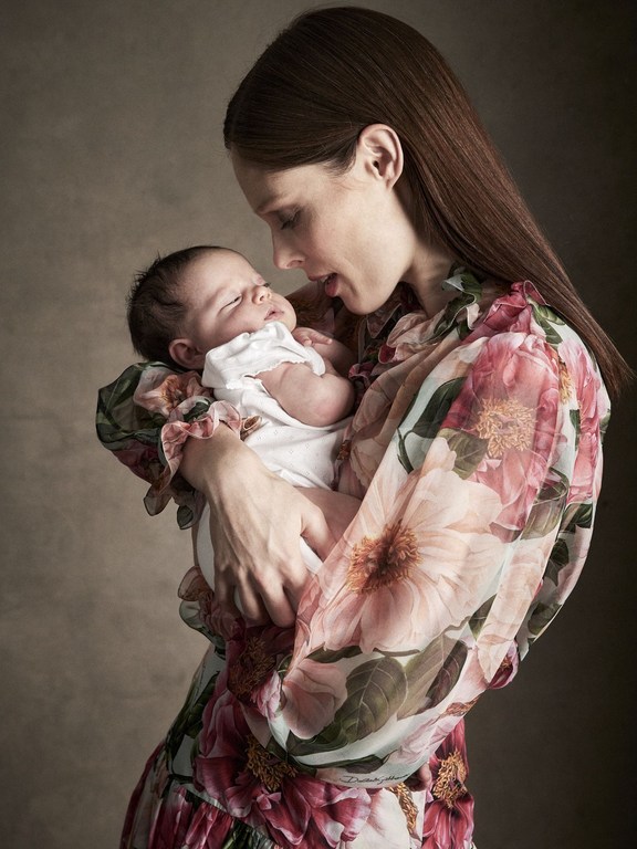 Mother holding newborn baby close in loving embrace, wearing a floral dress with a neutral background. Portrait.