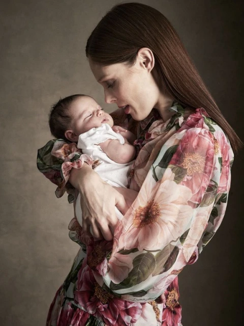Mother holding newborn baby close in loving embrace, wearing a floral dress with a neutral background. Portrait.