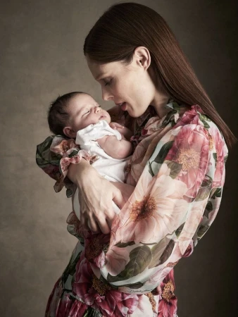 Mother holding newborn baby close in loving embrace, wearing a floral dress with a neutral background. Portrait.