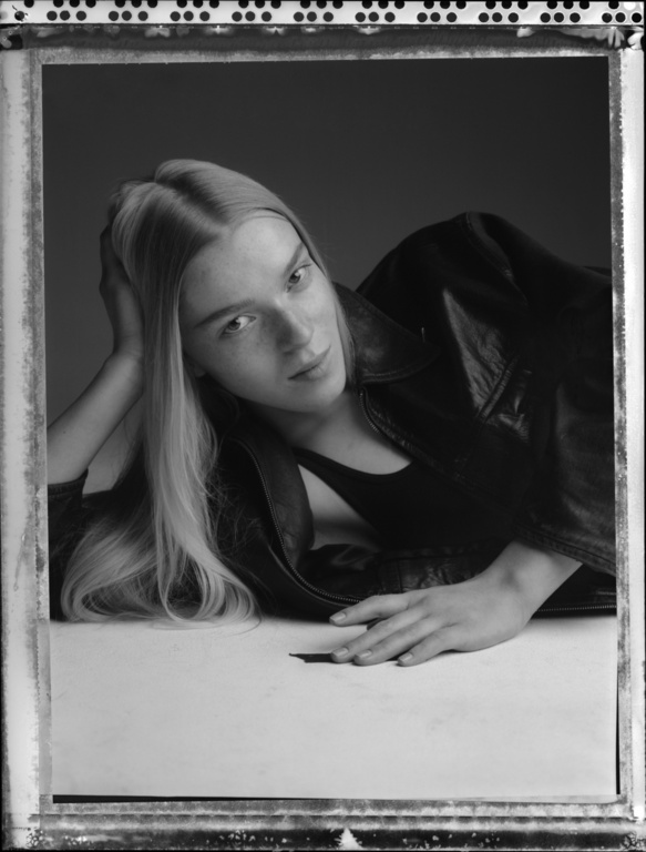 Monochrome portrait of a woman with long blonde hair wearing a dark jacket, resting on a light surface with a dark grey backdrop.