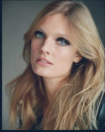 Close-up portrait of a blonde woman with blue eyes and dark eyeshadow wearing a black leather jacket against a grey background.
