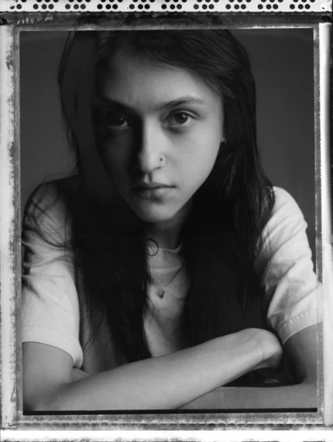 Monochrome portrait of a young woman with a nose ring and long dark hair. She is wearing a casual top and has her arms crossed.