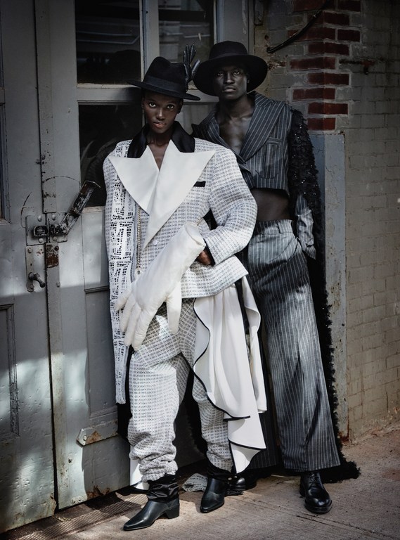 Two models pose outdoors wearing unique, patterned outfits with hats and boots in a fashion editorial-style portrait with urban background.