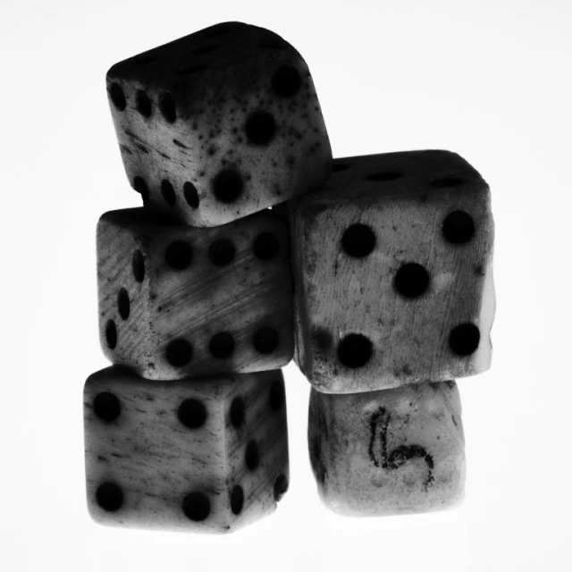 Monochrome photograph of a stack of five weathered dice, set against a bright white backdrop. The dice have black spots.