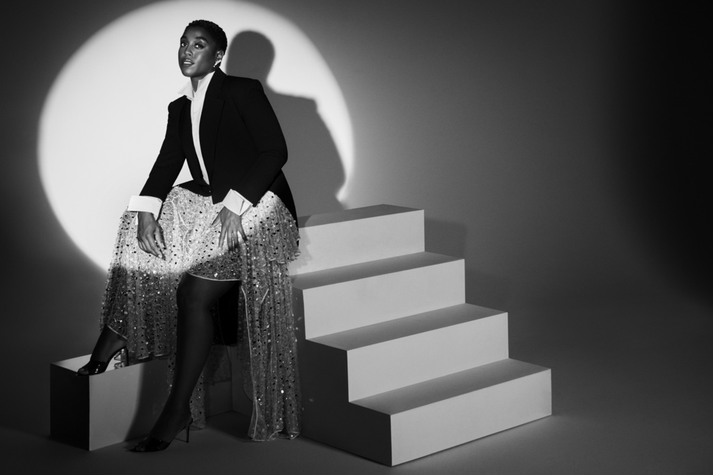 Black and white portrait of a woman in a blazer and sequined skirt sitting on white steps, bathed in the light of a spotlight.