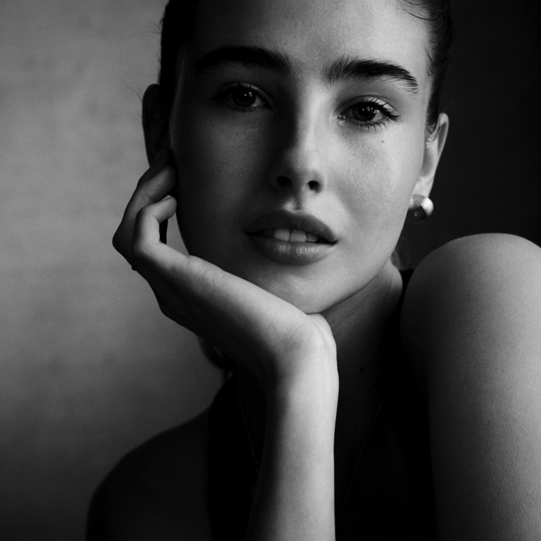 Monochromatic portrait of a young woman with hand on chin, wearing hoop earrings, against a blurred background in a close-up shot.
