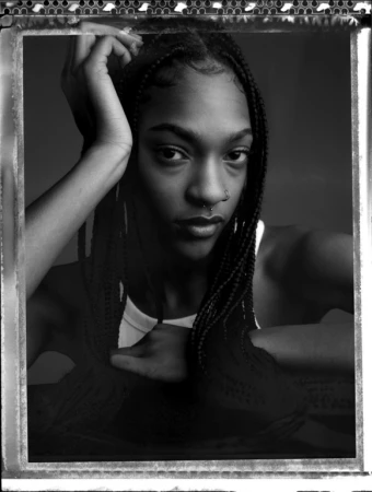 Black and white portrait of a woman with braided hair and nose ring resting her hand near her head, framed in a dark studio.