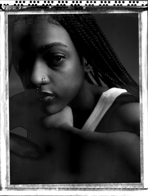 Monochrome portrait of an adult female with braided hair and nose rings, looking at the camera in a dark studio setting.