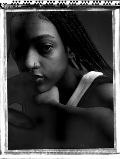 Monochrome portrait of an adult female with braided hair and nose rings, looking at the camera in a dark studio setting.