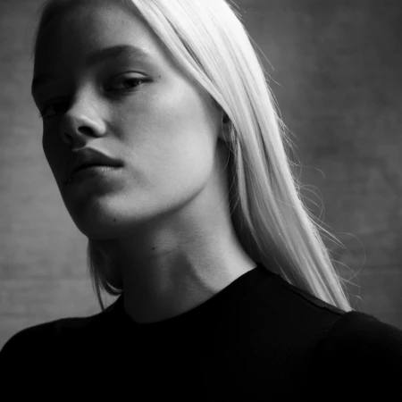 Close-up black and white portrait of a blonde woman wearing a black top against a textured wall background.