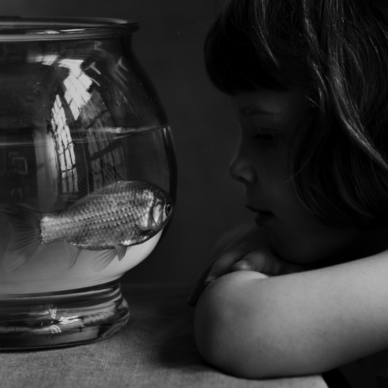 A black and white photo captures a young child gazing intently at a fish swimming in a round fishbowl, creating a simple, intimate scene.