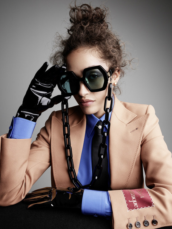 Model wearing oversized sunglasses, camel blazer, blue shirt, and black latex gloves photographed by Victor Demarchelier.