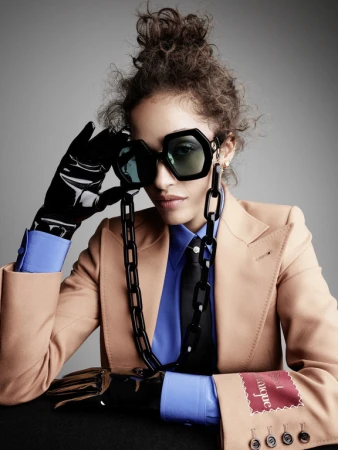 Model wearing oversized sunglasses, camel blazer, blue shirt, and black latex gloves photographed by Victor Demarchelier.