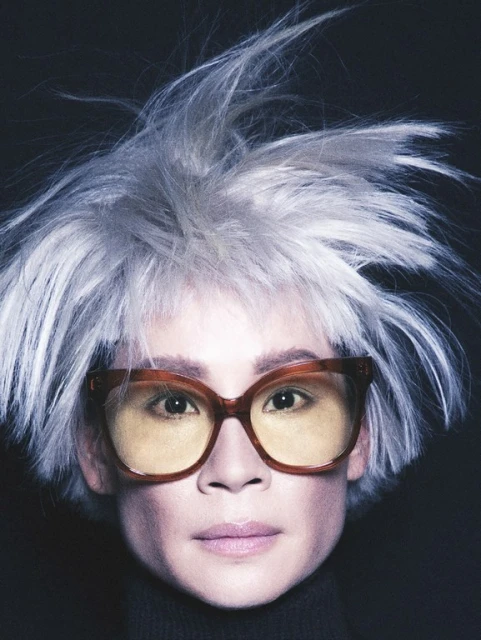 Close-up studio portrait of a person with white hair, wearing yellow-tinted glasses and a black turtleneck sweater against a dark backdrop.