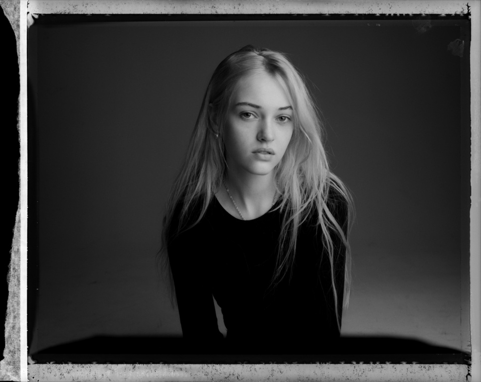 Portrait of a young blonde woman with long hair wearing a dark top against a plain studio background, photographed by Victor Demarchelier.